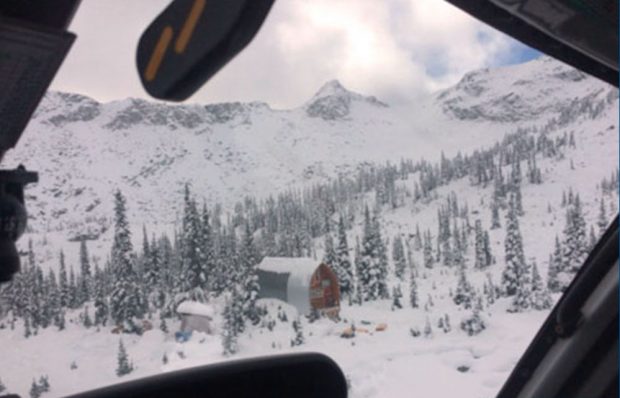 Vue aérienne du refuge depuis un hublot d’hélicoptère : de la neige couvre le sol, des arbres à feuilles persistantes ainsi que de hauts sommets situés derrière le refuge.