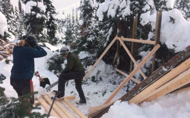 Le prédormant d’une nouvelle remise de bois a été érigé et un membre tenant une scie à chaîne s’apprête à tailler des montants de deux par quatre sur le sol tandis qu’un autre membre le regarde faire.