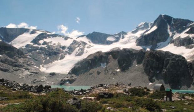 Le soleil estival tape dur sur les grands sommets glaciaires se dressant derrière l’eau bleu vert du lac contrastant avec le rocher gris foncé. Le refuge et les bécosses se trouvent plus loin à droite.