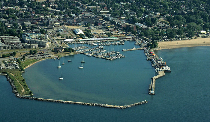 Une photo aérienne du port de Cobourg montre une longue jetée au bas qui rencontre presque la fin de l’autre quai situé à droite, et une ouverture étroite pour entrer dans le port. L’eau est bleue et, à l’intérieur du port, des voiliers sont ancrés ou amarrés à une marina. De gauche à droite sur la rive, des condos, de grandes tentes érigées pour le festival de Cobourg et des caravanes sont stationnées devant la plage. Il y a, au loin, des immeubles commerciaux, des résidences et des arbres feuillus.