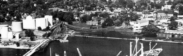 Une photo en noir et blanc du port industriel avec des réservoirs à pétrole blancs à gauche, des lignes de chemin de fer et des tas de charbon au centre ainsi qu’un wagon couvert à droite. Le Victoria Hall se trouve au loin à droite.