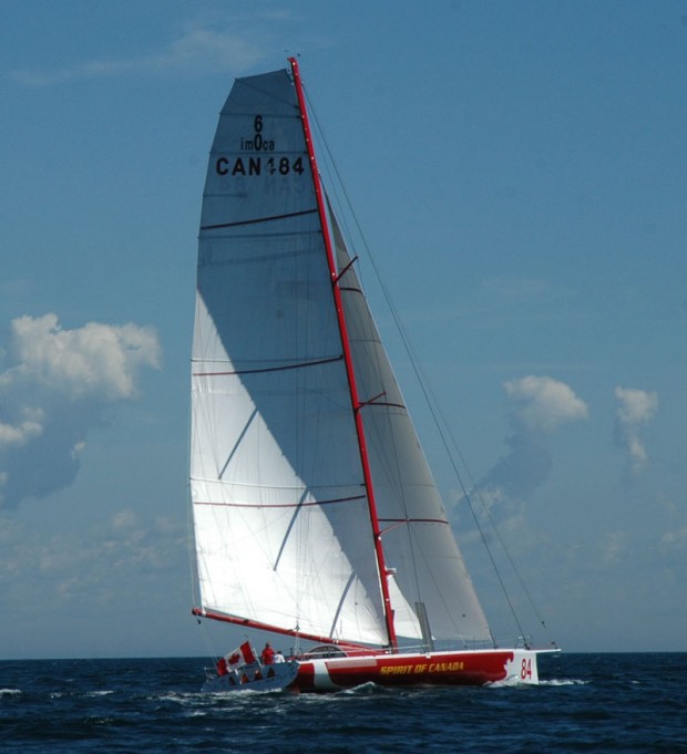 Une photo en couleurs d’un voilier aux lignes pures, avec une coque rouge et blanche, naviguant sur l’eau d’un bleu foncé, ses deux voiles blanches contrastant avec le ciel bleu. Un drapeau du Canada flotte à l’arrière du bateau.