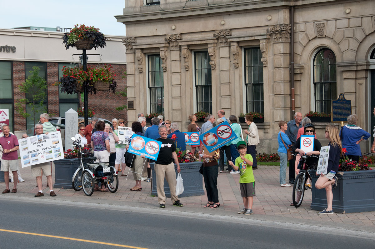 Une photo en couleurs d’une petite foule d’adultes et d’enfants brandissant des affiches sur le trottoir devant un immeuble.  