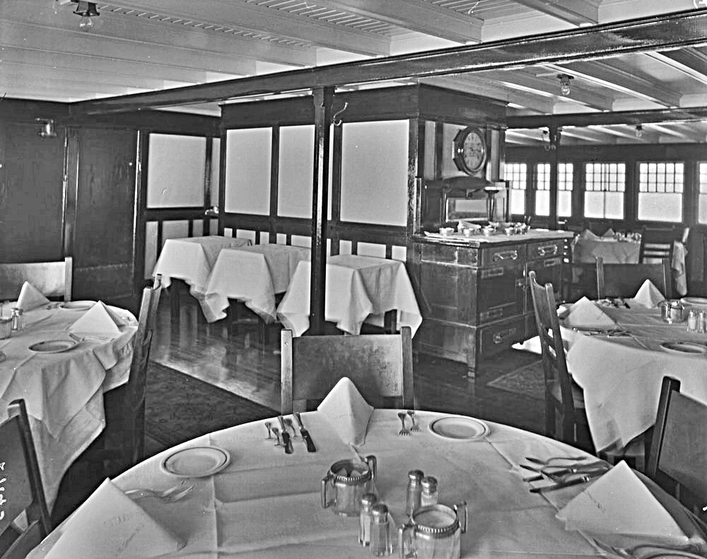 Photo en noir et blanc d’une vaste salle à manger avec des planchers de bois franc luisants et des tapis. Les tables rondes sont mises avec des nappes blanches, des serviettes de table, sept couverts. Les tables de service sont au centre en arrière-plan.  