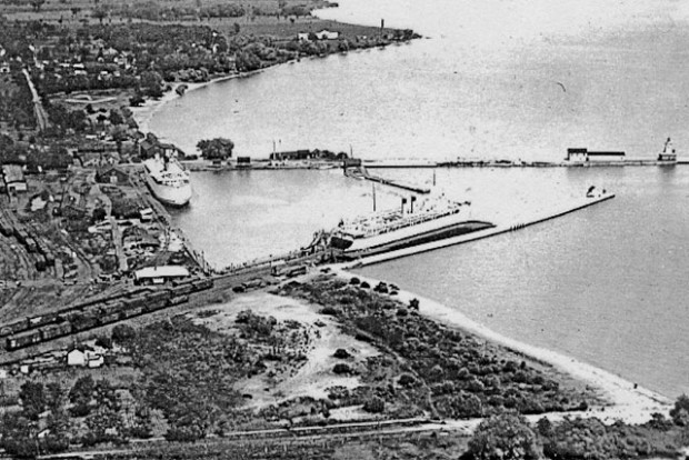 Une photo aérienne en noir et blanc montrant les deux transbordeurs amarrés au port de Cobourg. L’un d’eux, au centre de la photo, est amarré à un quai la proue face au lac et l’arrière collé à la fin d’une ligne de chemin de fer sur laquelle se trouvent de nombreux wagons. L’autre transbordeur est amarré à un quai le long de la rive. La photo inclut, en avant-plan, l’agrandissement du port du côté ouest et la plage plus loin.  