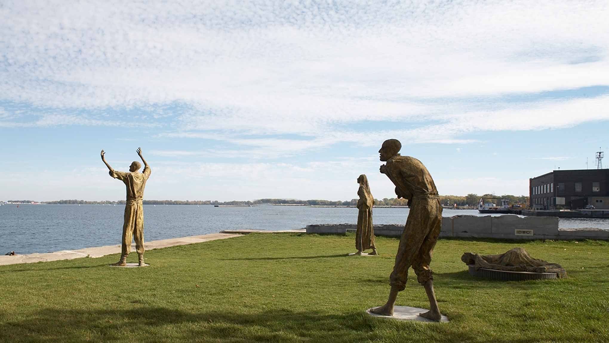 Quatre statues de bronze érigées sur une étendue de gazon près de la rive. Elles font toutes face au lac et il y a une certaine distance entre chacune. Une statue a les bras levés au ciel, une autre a le dos voûté, la dernière est étendue au sol.