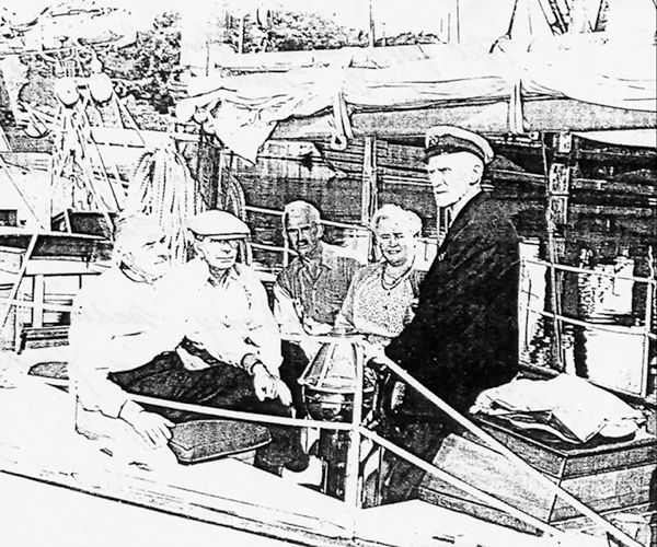 Une photocopie de qualité médiocre d’une photo de cinq personnes à l’arrière d’un bateau, quatre sont assis et le cinquième, qui porte une casquette de capitaine, est debout. 