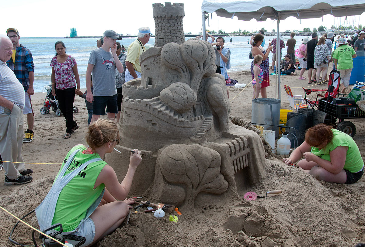 Deux sculptrices sur sable ajoutent la touche finale à un grand château au bord de l’eau bleue ; les passants s’arrêtent pour admirer leur travail.