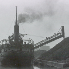 Une photo sombre en noir et blanc montrant, à gauche, un charbonnier vu de l’arrière avec une rampe descendant du vaisseau et versant le charbon pour former un énorme amoncellement sur le quai à droite.