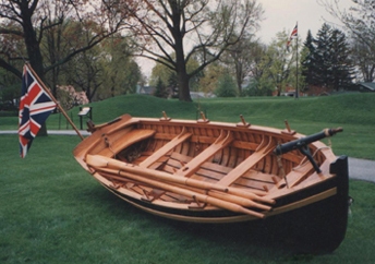 Petite embarcation de bois, avec un drapeau de l’Union Jack, repose légèrement inclinée sur l’herbe. Elle comprend quatre ensembles de dames et quatre bancs de rameurs, dont deux avec des trous pour les mâts.