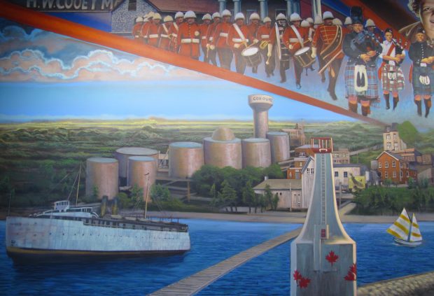 Une peinture murale aux multiples couleurs illustrant, à droite, le phare blanc actuel de Cobourg et, à gauche, un grand transbordeur, blanc lui aussi. Sur la rive, un peu en retrait vers la gauche, sept grands réservoirs à pétrole couleur argent et un réservoir d’eau surélevé. À droite, derrière le phare, il y a quelques entreprises. L’harmonie de Cobourg, habillée en uniforme rouge, et le corps de cornemuses de la légion, en kilts variés, défilant en formation militaire, sont dépeints au-dessus des nuages.