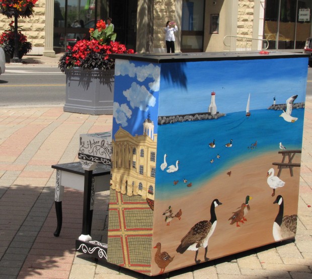 Photo en couleurs d’une scène de rue avec l’arrière et le côté d’un piano sur un trottoir pavé en avant-plan. Sur le côté du piano, on a peint le Victoria Hall sous un ciel bleu, et à l’arrière, il y a le port ensoleillé avec des oiseaux marins et des cygnes en avant-plan, et l’entrée du port avec le phare en arrière-plan. Un banc de piano est devant le piano, et, en bordure du trottoir, il y a un grand bac de fleurs rouges. Une personne de l’autre côté de la rue prend une photo.