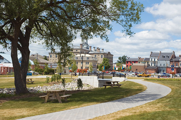 Une photo en couleurs d’un parc où on retrouve un grand arbre et plusieurs autres arbres plus petits, trois tables à pique-nique et un trottoir au milieu. En arrière-plan, il y a le Victoria Hall en brun et des immeubles commerciaux.