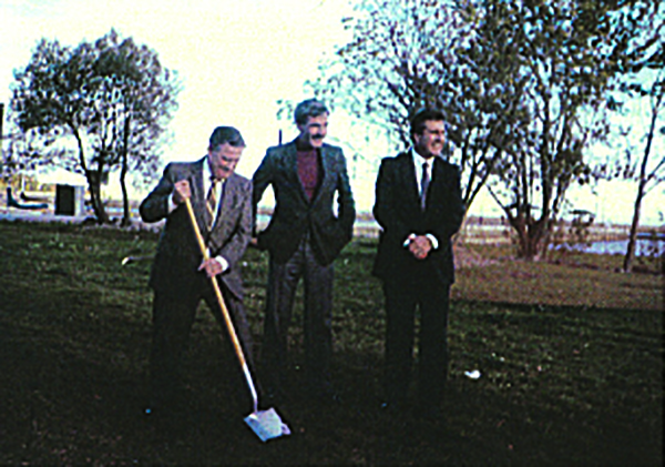 Une vieille photo en couleurs de trois hommes en habit qui sourient en posant sur une étendue de gazon vert avec la rive en arrière-plan. L’homme à la gauche pousse la pelle dans la terre.