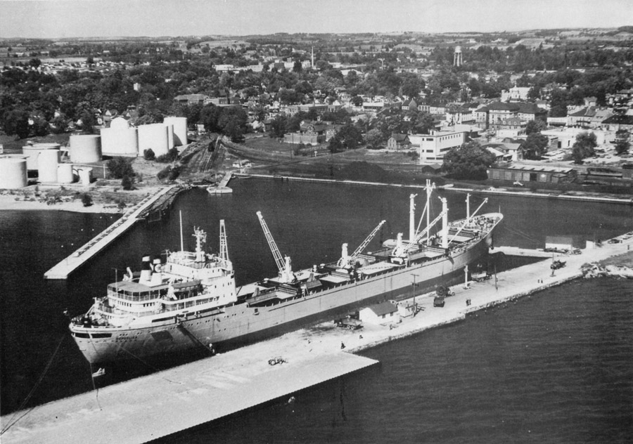 Une photo en noir et blanc d’un grand cargo, au milieu, amarré le long d’un quai avec deux grues à bord. Un deuxième quai mène aux réservoirs à pétrole sur la rive à gauche où se trouvent, entre deux quais, des amoncellements de charbon et un wagon le long de la rive. 