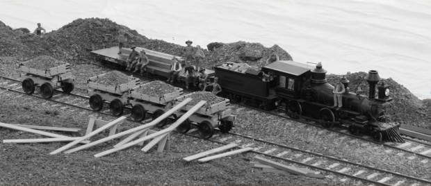 Une photo en noir et blanc du modèle réduit inspiré d’une photo rare. En avant-plan, il y a des madriers de bois appuyés contre un des quatre wagons de minerai sur la voie ferrée. Derrière, on retrouve une locomotive à vapeur auquel sont attachés un wagon rempli de bois et une remorque à plateau. Une douzaine de travailleurs dispersés posent pour la photo.