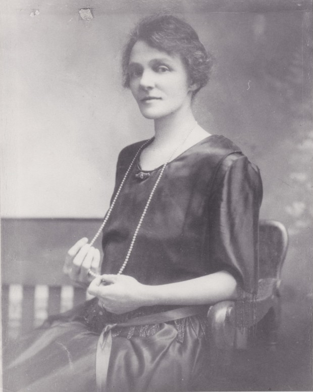 Violet Guymer en tant que jeune femme posant pour un portrait. Elle est vêtue d'une robe noire et a un collier de perles qui descend jusqu’aux genoux.