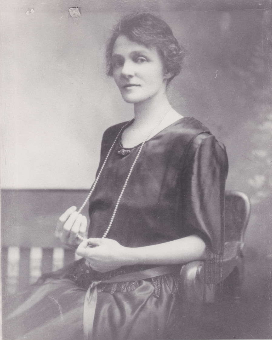 Violet Guymer en tant que jeune femme posant pour un portrait. Elle est vêtue d'une robe noire et a un collier de perles qui descend jusqu’aux genoux.