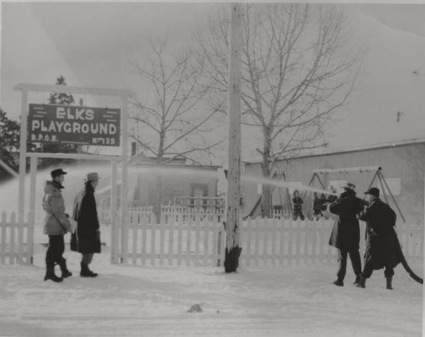 Il y a deux hommes qui ont un tuyau d’arrosage dans les mains et qui projettent de l’eau vers le terrain de jeu. On dirait qu'ils fabriquent une patinoire. En arrière-plan, il y a deux enfants qui jouent sur des balançoires.