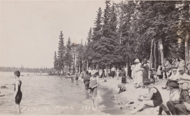 Des gens sont en maillots de bain avec plusieurs d'entre eux pataugeant dans l'eau. Il y a une petite plage, mais beaucoup d'arbres (principalement des conifères) en arrière-plan.