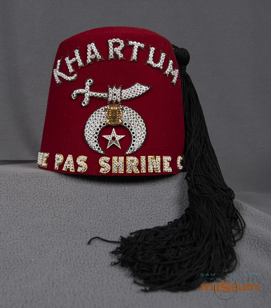 Un fez rouge des Shriners. Dessus, il y a quatre trous d’aération en forme de cercles et trois rangées de trous dans chaque cercle. « KHARTUM THE PAS SHRINE CLUB » est inscrit devant en lettres ornées de perles. Il y a aussi un symbole d’une épée arabe et un écusson égyptien avec une étoile argentée en-dessous. Un long gland noir est attaché en haut et pend sur le côté. Du côté droit, il y a une broche en argent avec une épée à clous arabe et une forme de croissant dessous; une feuille d'érable est au centre.