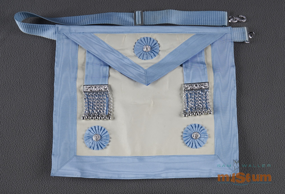 Tablier maçonnique avec une bordure bleue et une bretelle ressemblant à celle d’une sacoche. Un rabat triangulaire pend à 1/3 du haut et a une broche maçonnique argentée avec un motif bleu à fleurs autour. Le derrière est un morceau de tissu en coton bleu et le devant est en cuir blanc avec une bordure autour des côtés et du rabat. Deux bandes bleues en tissu pendent avec une barre argentée métallique et on peut voir sept chaînes et petits globes en métal qui pendent de chaque barre. Il y a des broches maçonniques dans les coins inférieurs. Il y a un serpent en forme de « S » sur la ceinture et une ceinture ajustable avec des fermoirs argentés en métal en haut sous le rabat.