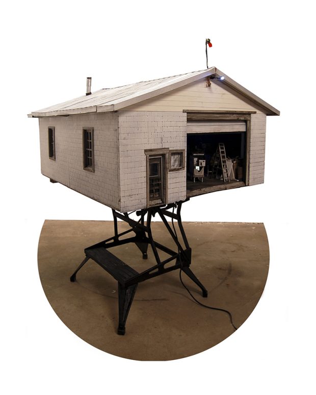 Vue du devant d’un atelier miniature avec un toit en métal et des briques de couleur grise qui sont posées sur une base en métal noire. La porte du garage est ouverte et à l’intérieur, on peut voir une échelle et plusieurs objets de l’atelier en miniature. 