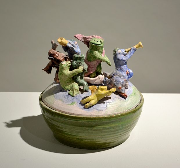 Un bol en céramique vert avec un couvercle. Sur le couvercle, plusieurs grenouilles et poissons, certains portant des habits, d’autres jouant du cor.