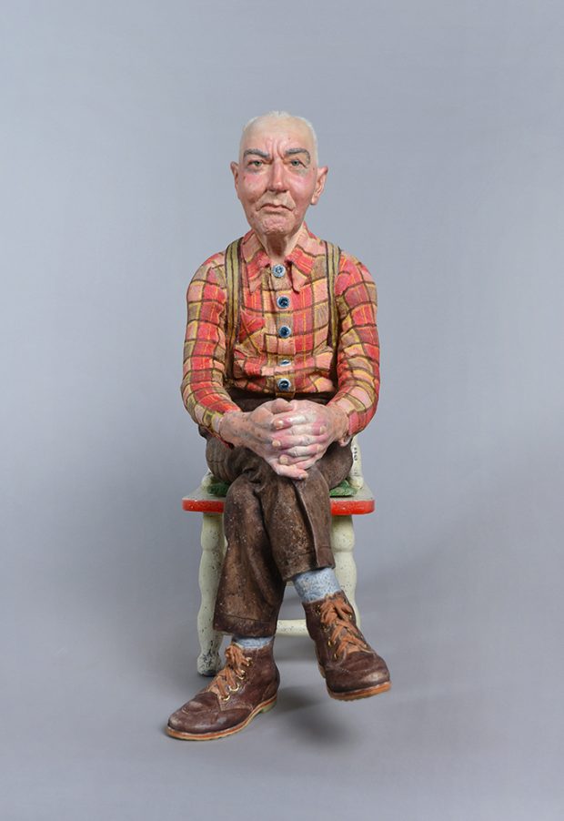 Sculpture représentant un homme assis qui porte une chemise à carreaux rouge et orange et un pantalon brun.