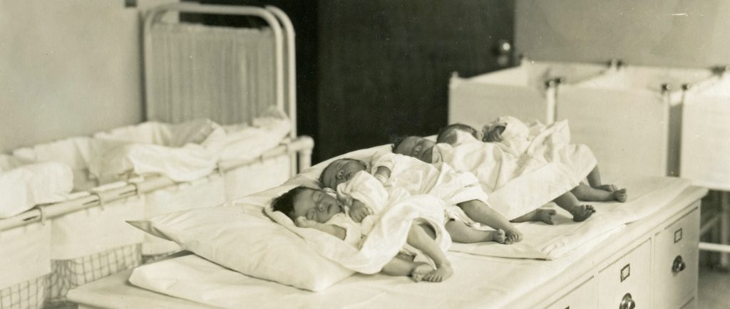 Nouveau-nés endormis alignés dans une rangée avec lits de bébé en toile de fond dans une photo en noir et blanc.