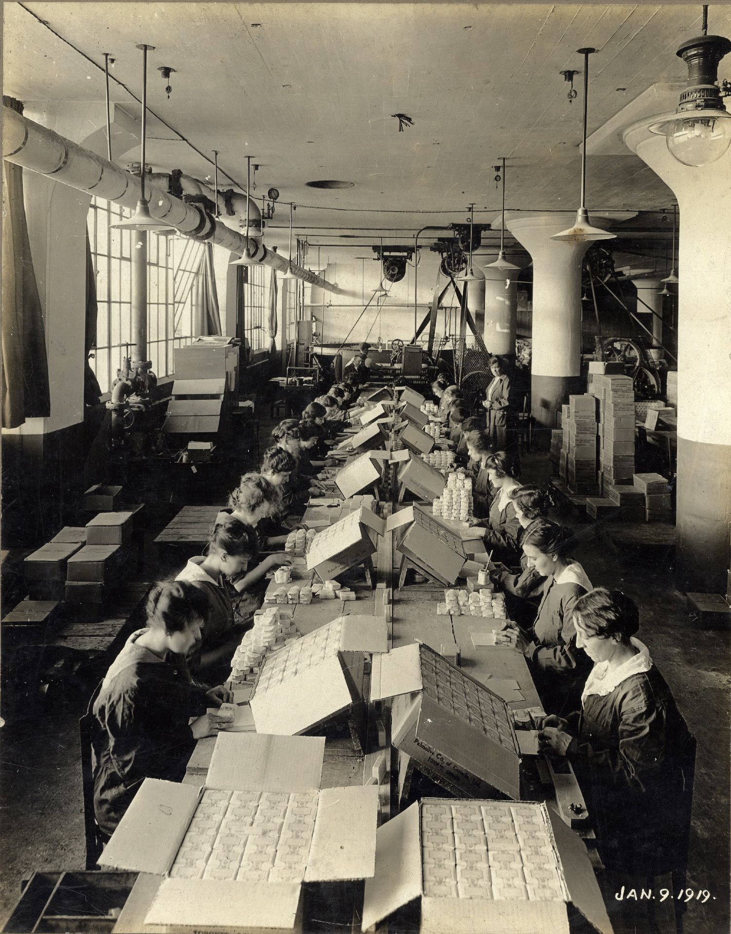 Deux rangées de femmes travaillent à une longue table dans un entrepôt dans une photo en noir et blanc. Des boîtes et équipements d’usine se trouvent en toile de fond.