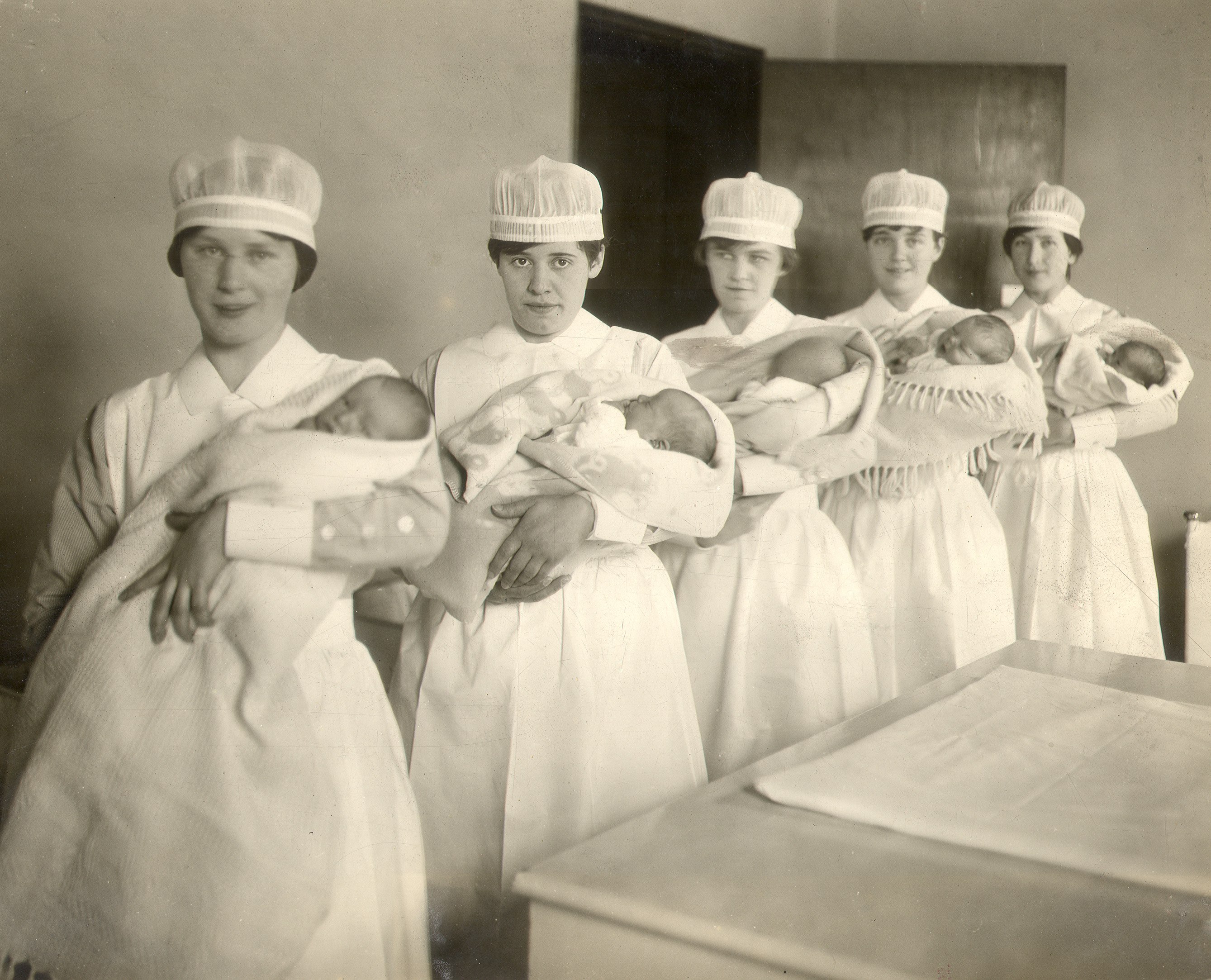 Cinq infirmières en uniforme tiennent des bébés enveloppés à côté d’une grande table dans une photo en noir et blanc.