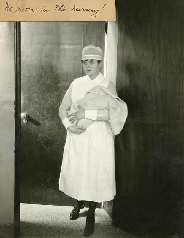 Une infirmière en uniforme tient un nouveau-né enveloppé dans une couverture pendant qu’elle sort d’une pièce dans une photo en noir et blanc.
