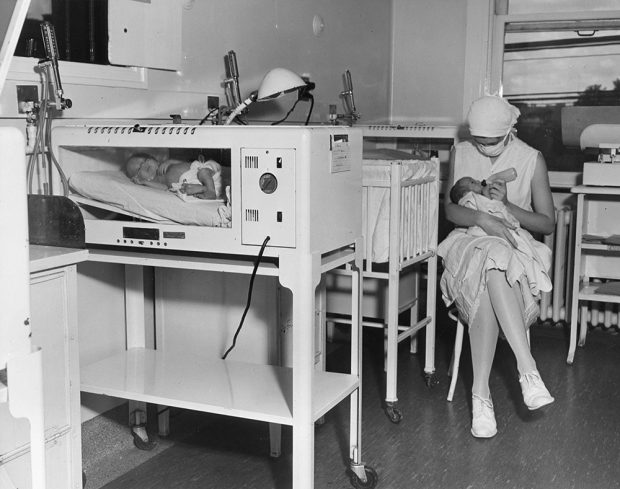 Une infirmière masquée nourrit un bébé assise sur une chaise à côté du lit de bébé dans une photo en noir et blanc. Un deuxième bébé se trouve dans un incubateur à l’avant-plan gauche.