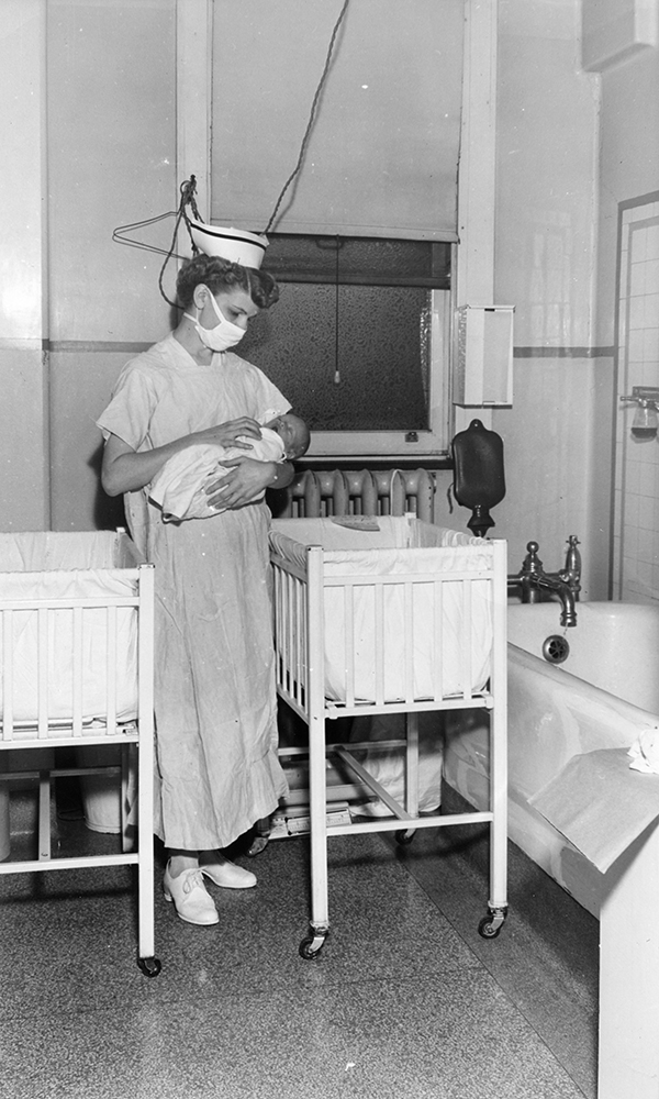 Une infirmière tient un bébé dans une petite pièce avec lits de bébé dans une photo en noir et blanc.