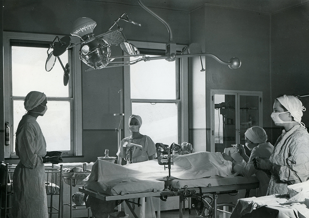Des employées médicales se tiennent autour d’un patient sur un lit dans une salle remplie d’équipement médical dans une photo en noir et blanc.