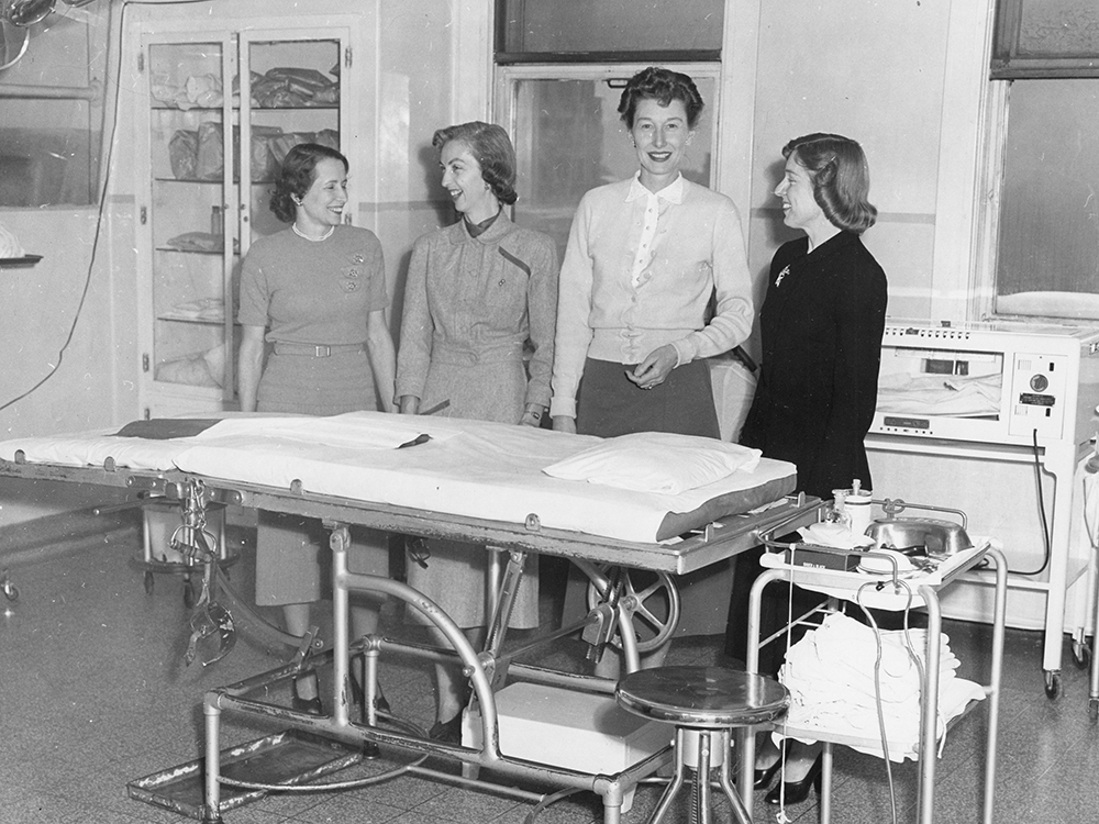 Quatre femmes habillées de vêtements de tous les jours se tiennent derrière une civière dans une salle avec de l’équipement médical dans une photo en noir et blanc.