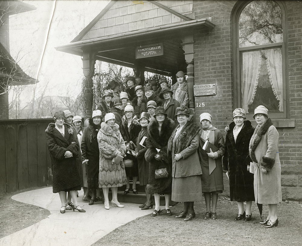 Un groupe de femmes habillées de manteaux d’hiver et de chapeaux à la mode à l’extérieur du Women's College Hospital dans une photo en noir et blanc.