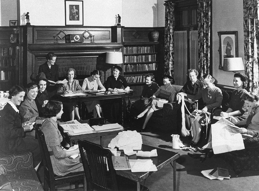 Des femmes assises dans un salon travaillent sur des documents et des tricots dans une photo en noir et blanc. Des tables ont été arrangées près des divans pour la réunion.