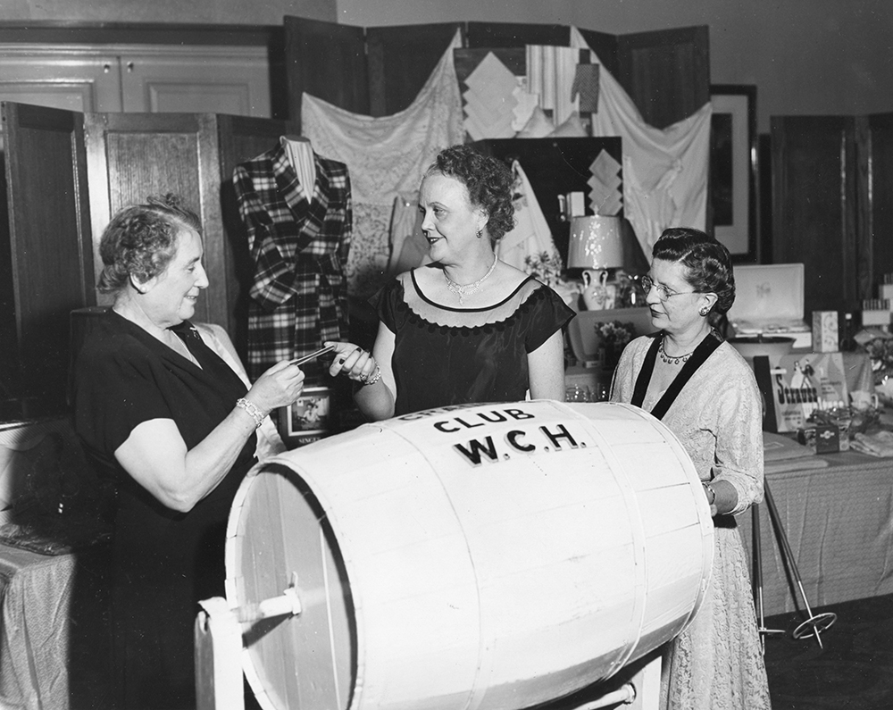 Trois femmes puisent un billet dans un baril étiqueté Cradle Club W.C.H. dans une photo en noir et blanc. Les objets à gagner sont étalés sur une table derrière elles.
