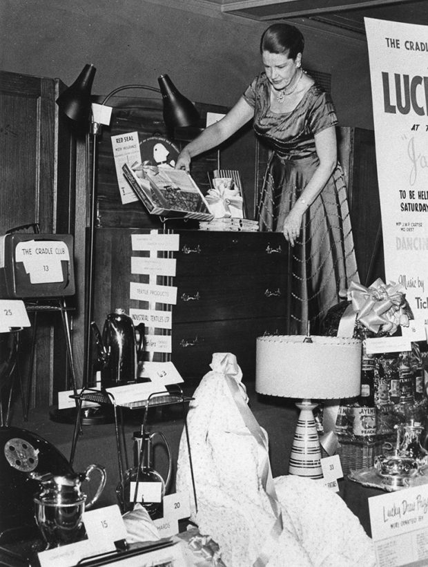Une femme en tenue formelle arrange des objets ménagers sur une scène dans une photo en noir et blanc.