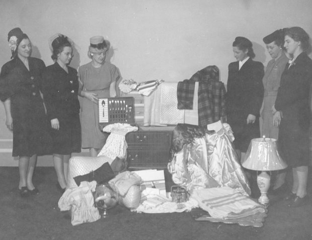 Six femmes en tenue formelle autour d’une collection d’objets ménagers et d’une grande commode dans une photo en noir et blanc. 