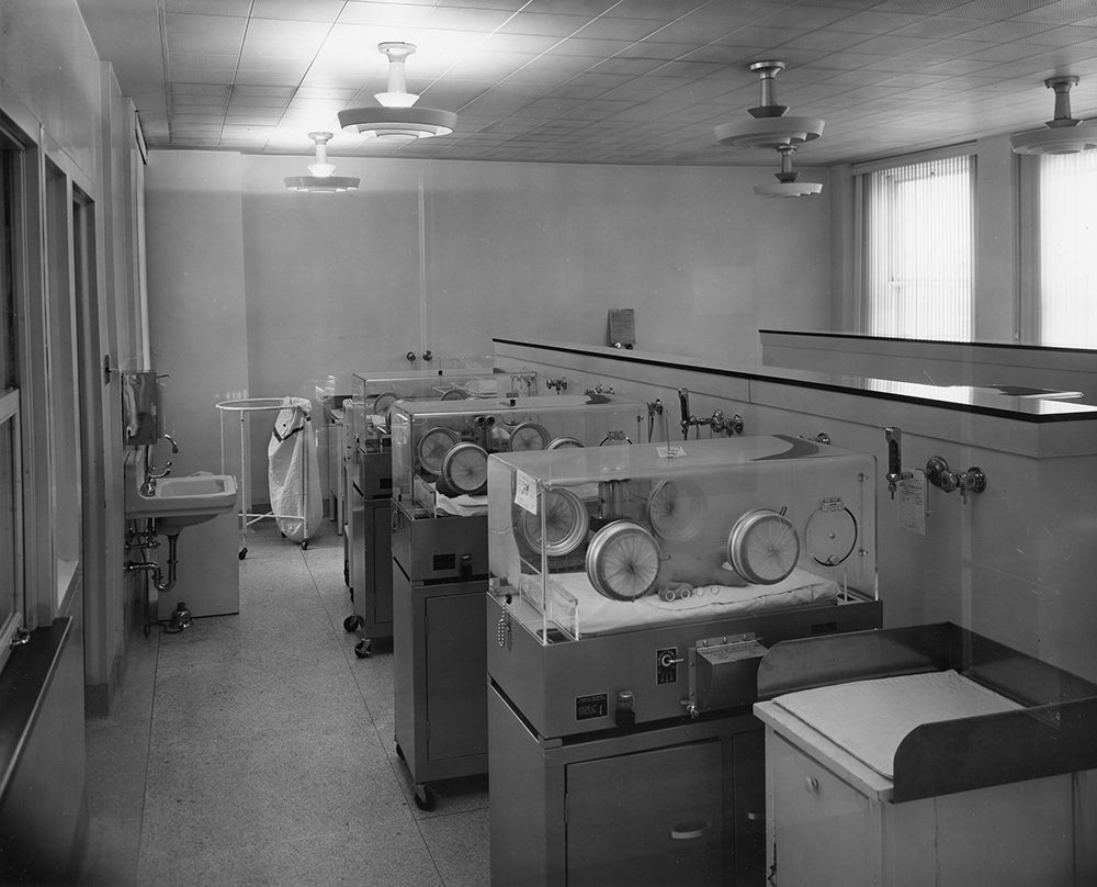 Une photo en noir et blanc d'un salle avec une rangée de trois incubateurs contenant des bébés. Un poste à langer se trouve à l’avant-plan et un lavabo à gauche.