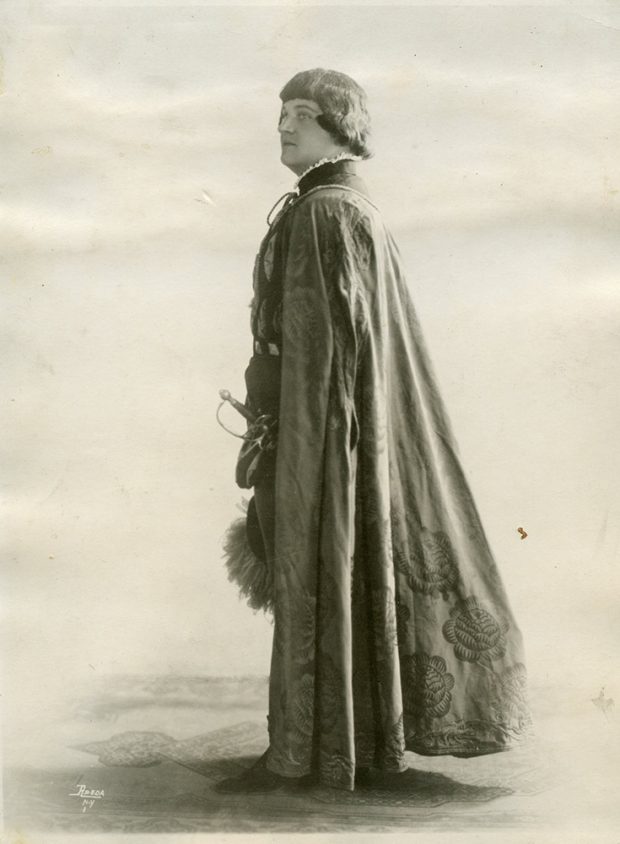 Homme vêtu d’un costume incluant cape et épée dans une photo en noir et blanc.