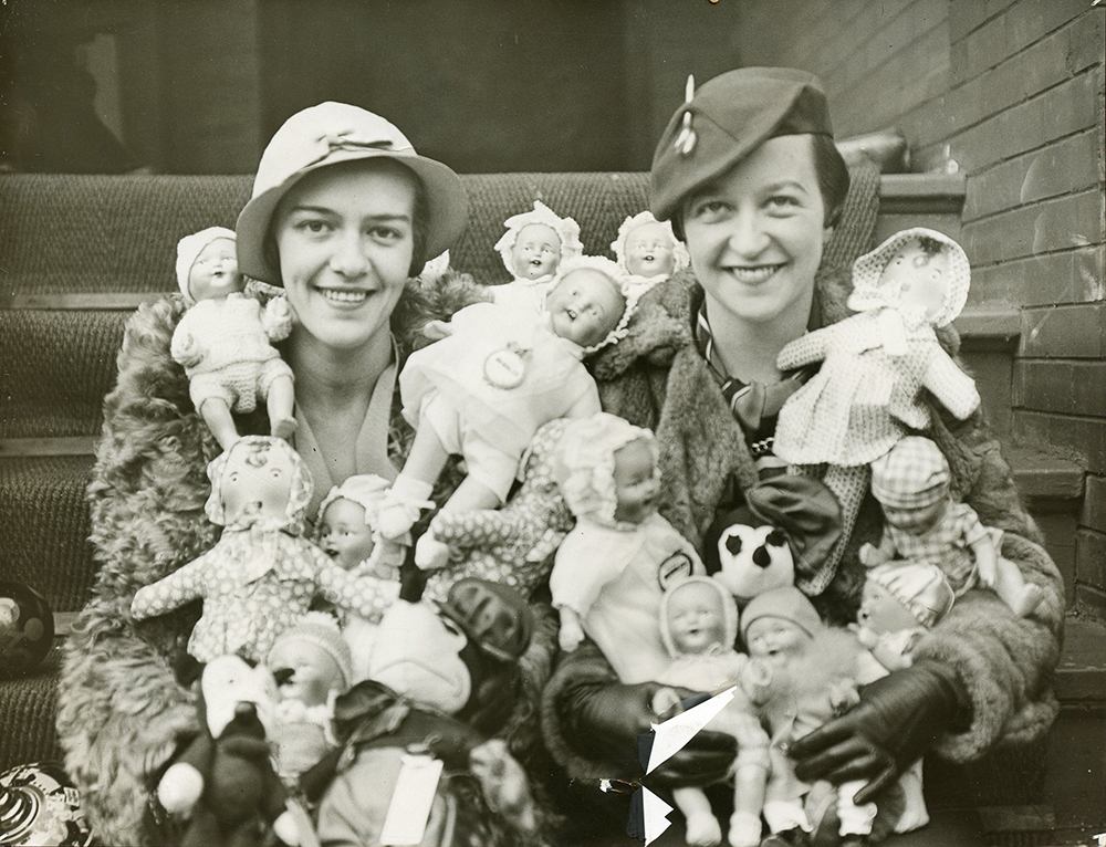 Deux femmes habillées à la mode s’assoient dehors sur un escalier les bras pleins de poupées dans une photo en noir et blanc.