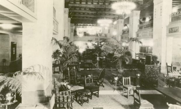 Hall d’entrée élégamment décoré avec choix de sièges divers, palmiers en pots et lustres dans une photo en noir et blanc.