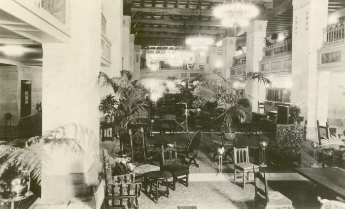 Hall d’entrée élégamment décoré avec choix de sièges divers, palmiers en pots et lustres dans une photo en noir et blanc.