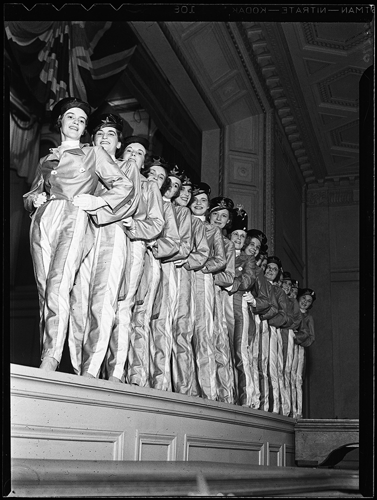Des femmes en costumes de fanfare identiques posent en rangée sur une scène dans une photo en noir et blanc. 