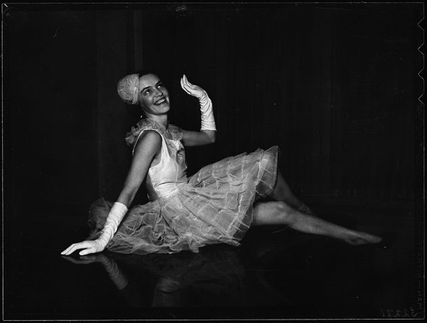 Peggy Beal assise à terre pose en tutu et gants dans une photo en noir et blanc.