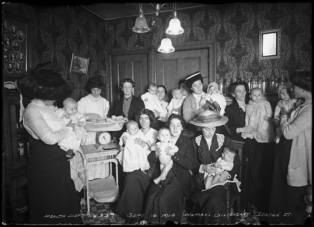 Un grand groupe de mères tenant leurs bébés posent dans un salon dans une photo en noir et blanc. Une infirmière pèse un bébé à gauche.